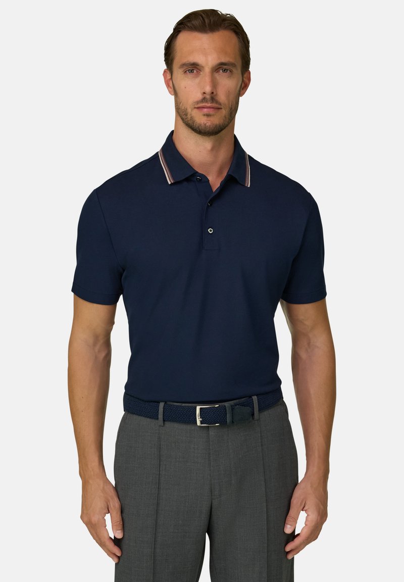 Homme portant un polo bleu marine avec un col rayé et un pantalon gris avec une ceinture tressée foncée, debout devant un fond uni.