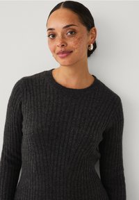 Frau mit dunklem Haar im Dutt, die silberne Creolen und einen schwarzen gerippten Pullover trägt, steht vor einer hellen Wand und lächelt sanft.