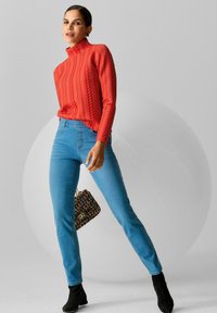 Rode gebreide trui met een hoge kraag, gecombineerd met blauwe slim-fit jeans en zwarte enkelboots. Een kleine meerkleurige handtas voegt detail toe.