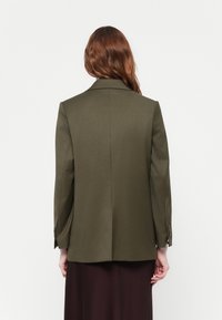 Olivově zelený tailored blazer s klasickým klopovým límcem, jedním větracím otvorem a dlouhými rukávy. Obsahuje jemnou texturu a zapínání na knoflíky u manžet.