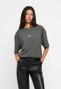Hiilenharmaa oversize t-paita, jossa on lyhyet hihat ja pyöreä kaula-aukko. Rinnassa on pieni valkoinen logo, ja se on valmistettu pehmeästä puuvillakankaasta.