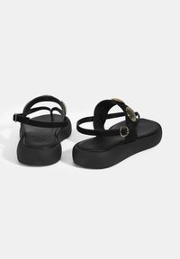 Schwarze Flip-Flops mit Plattform-Design, ausgestattet mit Wildlederriemen und runden goldenen Akzenten. Texturierter Fußbereich mit verstellbarem Knöchelriemen.