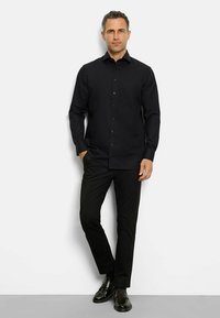 Camicia nera con bottoni, maniche lunghe, tasca sul petto e colletto rigido. Abbinata a pantaloni neri e scarpe nere lucidate.