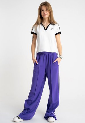 Młoda kobieta stojąca z rękami w kieszeniach, ubrana w białą koszulkę polo z czarnym obszyciem i luźne fioletowe spodnie na jasnym jednolitym tle.