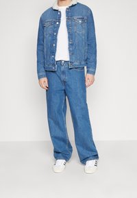 Man med blå denimjacka med vit fleecekrage, vit t-shirt, vida blå jeans och vita sneakers med svarta ränder.