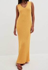 Une longue robe jaune en tricot côtelé avec un décolleté en V et un design sans manches, associée à des sandales marron à brides. Texture lisse.