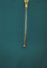 Türkisfarbener Stoff mit glatter Textur und einem goldenen Reißverschluss in der Mitte, der die Zähne und den Zuggriff des Reißverschlusses hervorhebt.