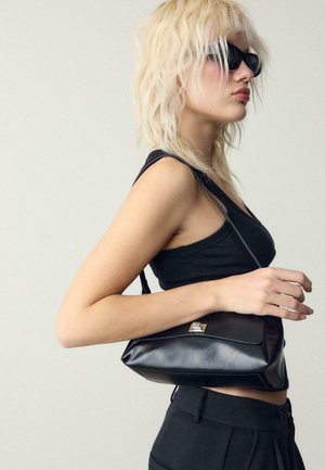 Blonde Frau, die ein schwarzes Tanktop und eine schwarze Hose trägt, schwarze Sonnenbrille aufhat und eine kleine schwarze Leder-Umhängetasche bei sich trägt.