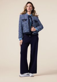 Giacca in tweed blu con due tasche sul petto, abbinata a un pantalone a gamba larga color blu scuro e sneakers grigio chiaro. Texture liscia, design cropped.