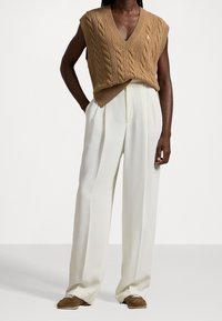 Knit sleeveless sweater in camel with cable pattern, v-neck, layered over cream wide-leg trousers. Brown slip-on shoes complete the outfit.