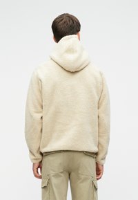 Άνδρας που στέκεται με την πλάτη στραμμένη, φοράει μπεζ φλις hoodie από sherpa και ανοιχτό καφέ cargo παντελόνι, με φόντο λευκό τοίχο.