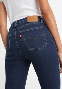 Dunkelblaue Jeans aus Denim mit Gesäßtaschen, versehen mit einem Leder-Label und gelben Nähten. Das Slim-Fit-Design betont die Figur.