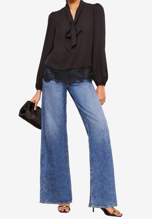 Frau trägt schwarze Langarmbluse mit Spitzenabschluss, weit geschnittene blaue Jeans, hält eine schwarze Clutch und trägt offene Sandaletten mit Absatz.