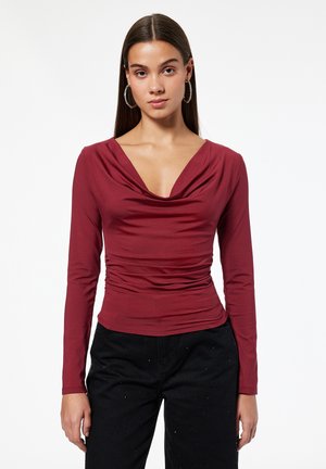 Kobieta o długich ciemnych włosach, nosząca duże kolczyki koła, dopasowaną bordową bluzkę z długim rękawem i marszczonym dekoltem oraz czarne spodnie.