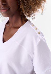 Witte katoenen T-shirt met een V-hals, voorzien van drie gouden knopen op de schouder en ribgebreide halslijn en mouwranden. Glad textuur.
