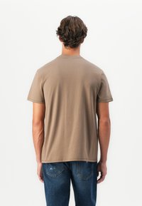 Hombre con cabello marrón ondulado que lleva una camiseta básica de manga corta marrón y jeans azules, de espaldas contra un fondo blanco.