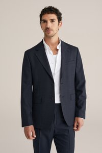 Marineblauwe wollen blazer met een inkeping in de revers, twee knopen en zijzakken. Gecombineerd met een wit overhemd en een donkere broek. Subtiele textuur en strakke lijnen.