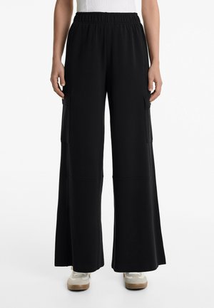 Pantalons larges noirs avec une taille élastique, dotés de poches latérales et d'une texture lisse. Associés à des baskets de couleur claire.