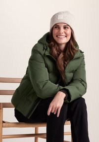 Green puffer jacket with a high collar and hood. Ribbed cream knit hat. Model seated on a wooden chair, wearing black pants.