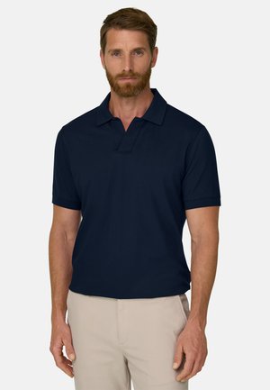 Homme barbu portant un polo bleu marine à manches courtes et un pantalon beige clair, debout devant un fond clair uni.