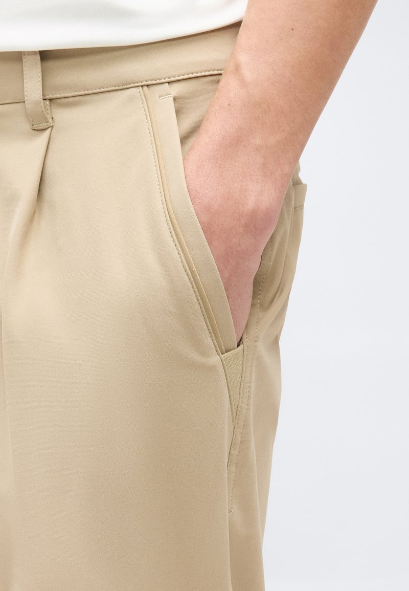 Main reposant à l'intérieur de la poche avant d'un pantalon beige, montrant la texture du tissu et le détail de la poche.