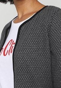 Persona che indossa una camicia bianca con lettering rosso sotto a un cardigan grigio con texture e bordi neri lungo il colletto e il davanti.