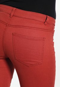 Czerwone dżinsy o gładkiej teksturze, z jedną tylną kieszenią i dopasowanym krojem, który podkreśla linię talii.