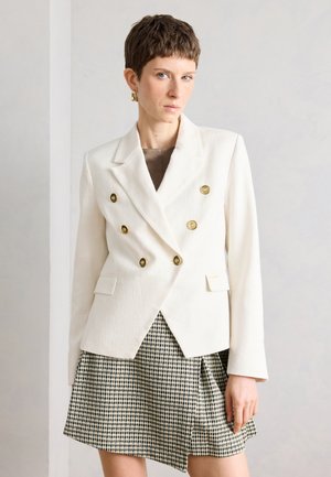 Kobieta o krótkich brązowych włosach nosi biały dwurzędowy blezer z złotymi guzikami oraz plissetowaną spódnicę w czarno-biały wzór pepitki, stoi w pomieszczeniu.