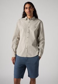 Chemise à manches longues beige clair, boutonnée avec un logo discret, associée à un short bleu. La chemise a une texture lisse et un design de col classique.