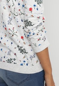 Sudadera de ligero gris con patrón floral, que presenta flores coloridas en rojo, rosa, azul y verde. Los puños y el dobladillo acanalados aportan textura.