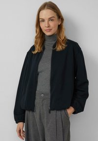 Czarna kurtka bomberka z zamkiem z przodu, elastycznymi mankietami i luźnym krojem, noszona na szarym golfie. Widoczne teksturowane tkaniny i boczne kieszenie.