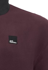 Burgundy fleece pullover with a black collar and a square black patch featuring the white logo "Jack Wolfskin." Soft texture and simple design.