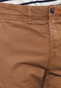 Brązowe chinosy wykonane z bawełny. Posiadają zapięcie na guzik, szlufki na pasek oraz kieszeń. Gładka struktura z delikatnym diagonalnym splotem.