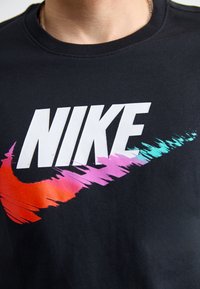 Črna bombažna majica s kratkimi rokavi, ki vsebuje velik bel logotip "NIKE" z gradientnim črtnim vzorcem v rdeči, roza in turkizni barvi.
