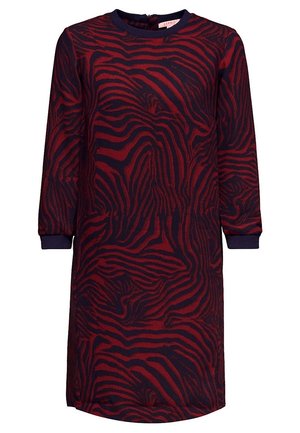 Femme portant un manteau noir arrivant aux genoux, par-dessus une robe rouge et noire à motifs, avec des bottines bordeaux à lacets, debout devant un fond gris uni.