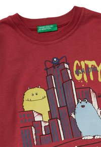 Rode T-shirt met cartoonmonsters en gebouwen. Het ontwerp bevat gele en blauwe monsters met de grafische tekst "CITY NEW YORK." Zachte katoenen stof.