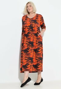 Maxi-Kleid in lebhaftem Rot und schwarzem abstraktem Muster, mit langen Ärmeln, einem runden Ausschnitt, Empire-Taille und seitlichen Taschen. Flaue schwarze Schuhe.