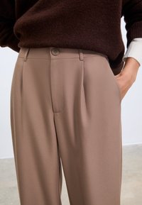 Beige maßgeschneiderte Hose aus glattem Stoff, mit vorderen Falten und einem Knopfverschluss. Kombiniert mit einem braunen Pullover, Hände in den Taschen.