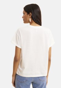Camiseta de algodón blanca con mangas cortas y cuello redondo. La parte trasera presenta un ajuste relajado, textura suave y sin patrones visibles.