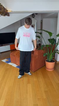 T-shirt branca oversized com logo "adidas" rosa, combinada com calças cargo pretas e sapatos prateados, em pé numa sala com plantas e mobiliário.