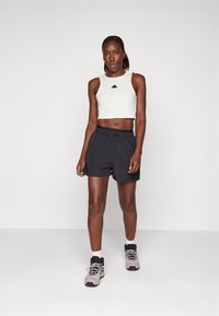 Fehér, cropped tank top fekete logóval, fekete rövidnadrággal párosítva. A modellen világoslila és fekete sportcipő van fehér zoknival. Egyszerű dizájn.