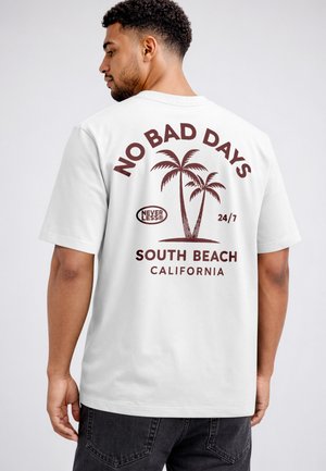 Mann trägt weißes T-Shirt mit braunem Palmenmotiv und Text „No Bad Days, South Beach California“ auf dem Rücken.