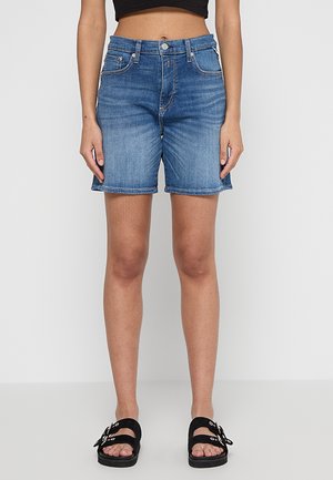 Jeansshorts in mittlerem Blau mit hoher Taille. Roher Saum und Fünf-Taschen-Stil. Kombiniert mit schwarzen Sandalen mit dekorativen Schnallen.