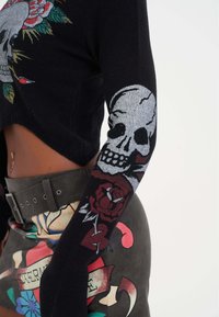 Maglione corto nero con ricami di teschi e fiori sulle maniche; abbinato a una gonna di pelle a vita alta con stampe colorate in stile tattoo.