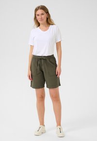 T-shirt blanc à col en V associé à un short en olive avec cordon de serrage, comportant deux poches avant et une coupe ample. Porté avec des baskets de couleur crème.