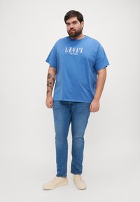 Mėlynas medvilnės marškinėliai su "LEVI'S" logotipu, laisvas kirpimas. Sujungti su vidutinio plovimo džinsais ir smėlio spalvos sportbačiais. Paprastas, kasdieniškas aprangos derinys.