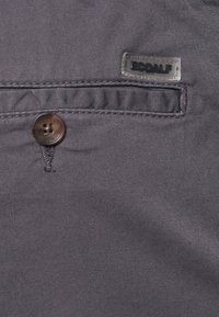 Mörkgrått tyg med en slät textur, prydd med en rund brun knapp och en genomskinlig Ecoalf-märkning på ett ficka.