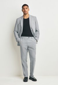 Abito grigio chiaro con rever a lancia, chiusura con bottoni e pantaloni a patta piatta. Abbinato a una camicia nera e scarpe nere lucide.