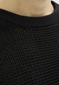 Black knitted sweater featuring a textured open weave design and a ribbed crew neckline. Textured surface with evenly spaced holes.