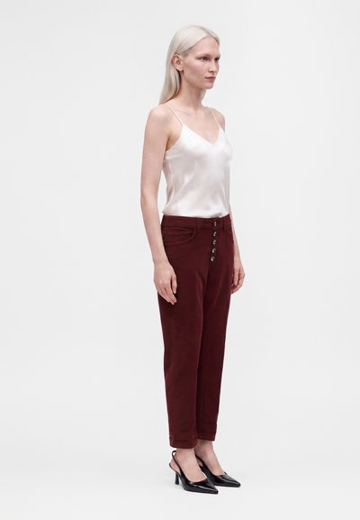 Femme aux longs cheveux blonds portant un caraco en satin blanc, un pantalon bordeaux boutonné et des talons noirs pointus, debout sur un fond blanc.